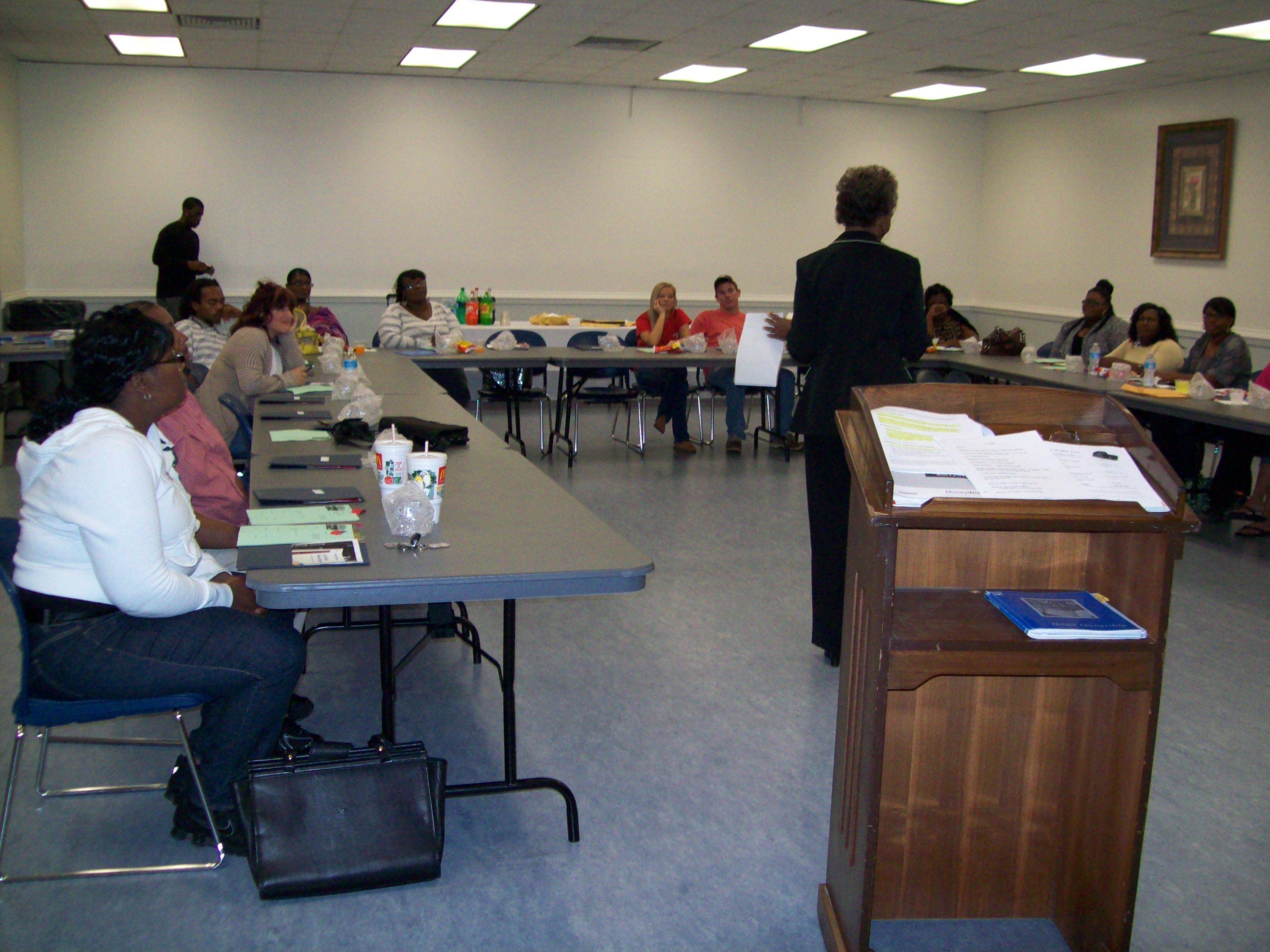 A photograph of approximately thirteen residents sitting in a meeting room at tables arranged in a u-shape. A counselor stands, with her back to the camera, at the open end of the table arrangement. Behind the counselor is a lectern with books and papers on it. 
