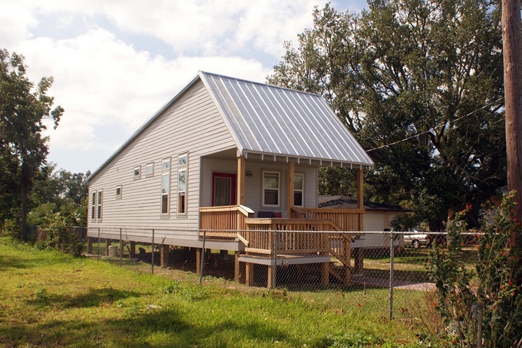 A photograph of a narrow but deep one-story single-family detached house. The wood-sided house, raised on wood pilings about four feet off the ground, has a porch across the front façade.