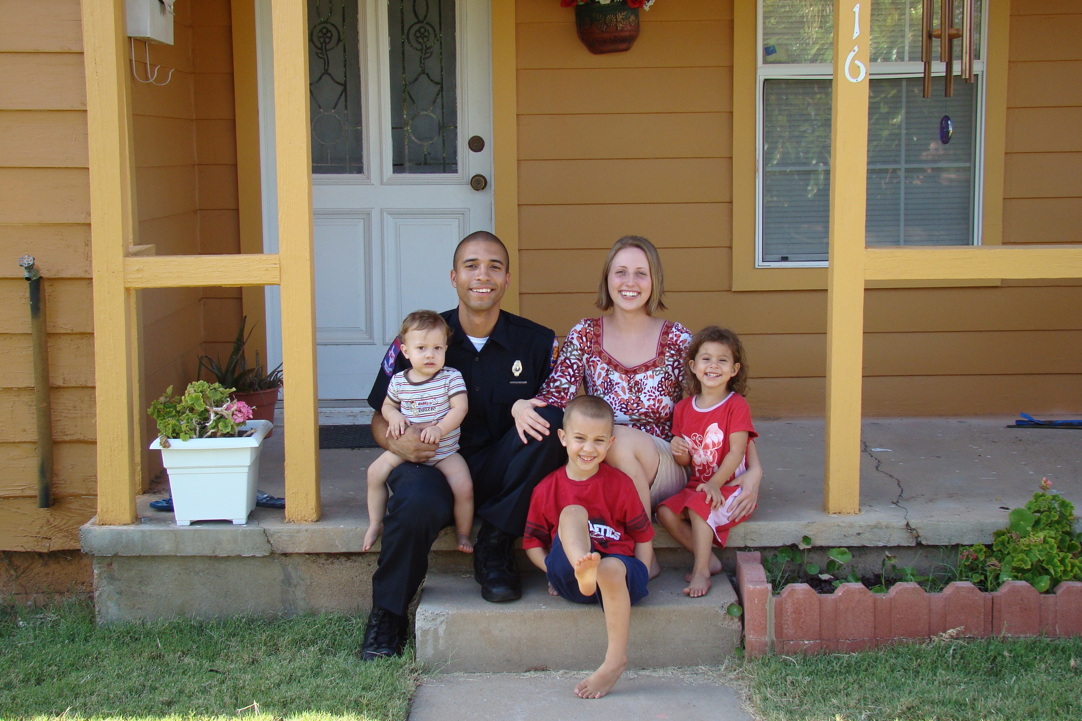 A photograph of a family of five, including two preschoolers and one toddler, sitting on the front porch step of their home.