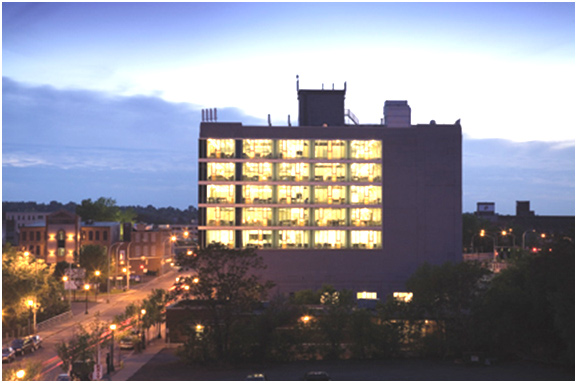 A photograph of the Nancy Cantor Warehouse in the evening. Most of the building is in shadows, with lighted windows on the top five floors of the seven-story building in the middle-ground of the photograph. To the left, in front of the warehouse building, is the Connective Corridor.