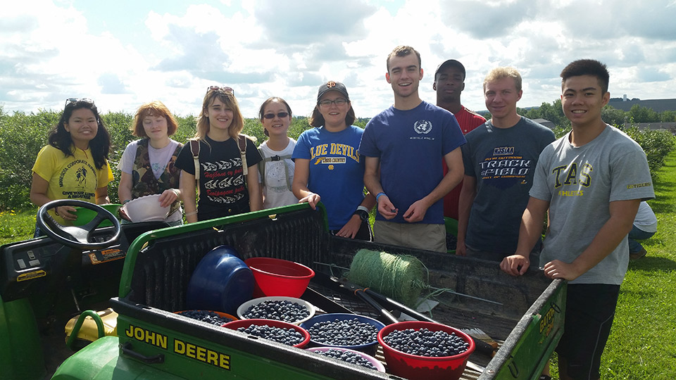 Photograph of nine Bowdoin students standing beside a utility vehicle loaded with several buckets of blueberries.