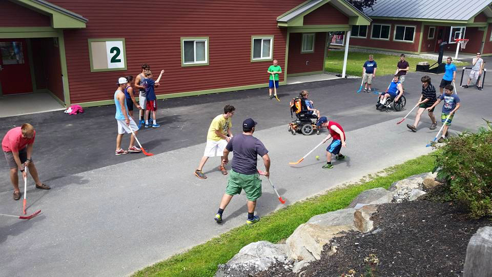 Photograph of 17 youth watching and playing field hockey in a paved area in front of a camp building.