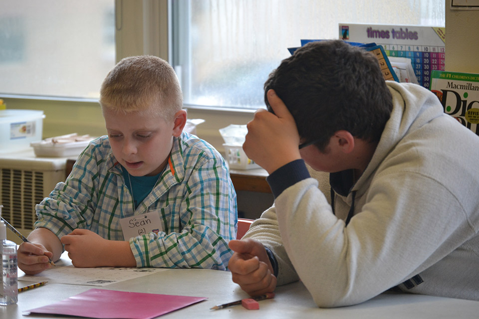 Photograph of a college student and a second-grade student sitting at a table in a classroom and discussing a worksheet.