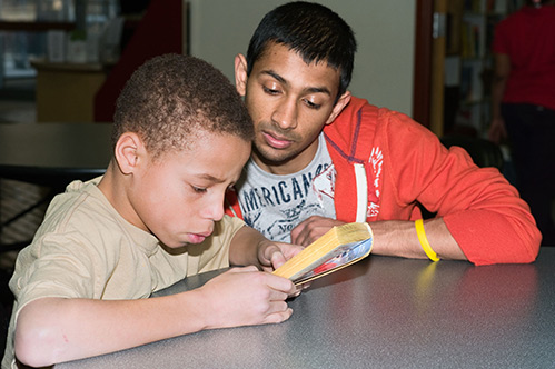 Photograph of a child and college student seated at a table; the child is reading a book while the college student follows along.