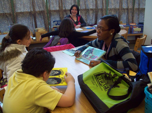 A woman holding a book sits at a table in a classroom with two children.